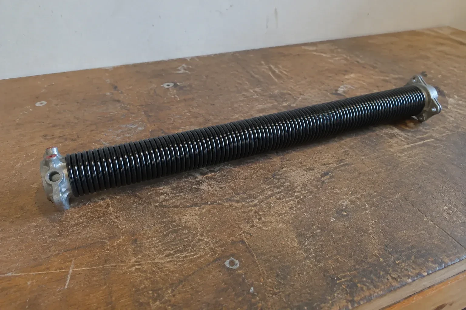 Detached black coil spring with metal end fittings on a workbench. — Torsion Spring Replacement, Lincoln